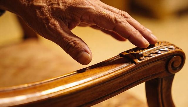 Senior hand touching ornate carved wood armrest on antique chair warm amber tactile detail