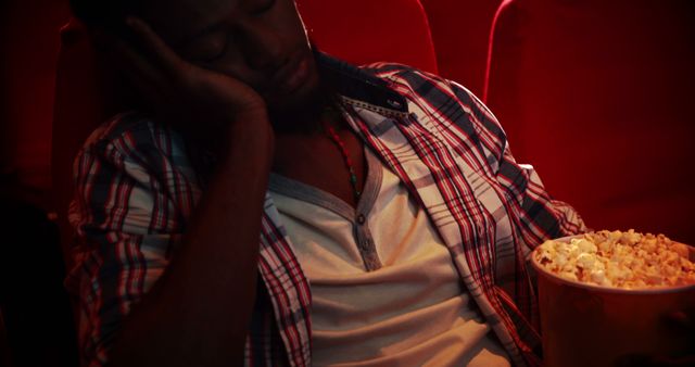 A young man is sleeping in a dimly lit movie theater while holding a bucket of popcorn. The plaid shirt and casual attire suggest he might be at a late-night screening or exhausted from a long day. This image can be used to depict themes of exhaustion, relaxation, or boredom in different scenarios. Perfect for blogs about cinema experiences, lifestyle content, or humorous social media posts.