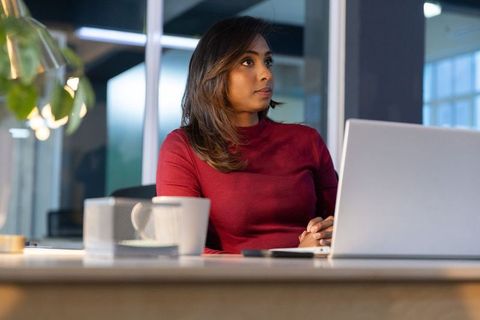 Indian professional woman working late on laptop in modern glass office holding coffee mug
