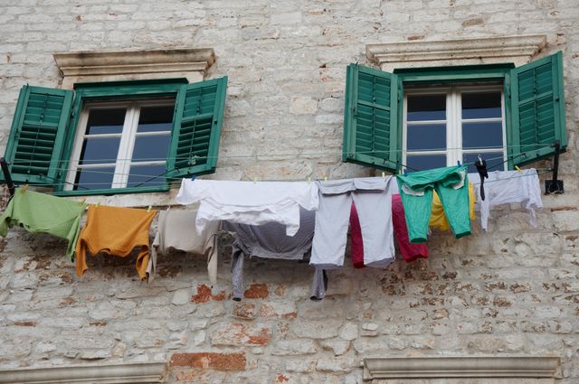 This image depicts colorful clothes hanging on a clothesline outside a historic stone building with green shuttered windows. The rustic appearance of the building, combined with the vibrant colors of the clothes, creates a visually appealing contrast. Perfect for use in travel blogs, articles about traditional lifestyles, or advertisements for rural tourism and outdoor activities.