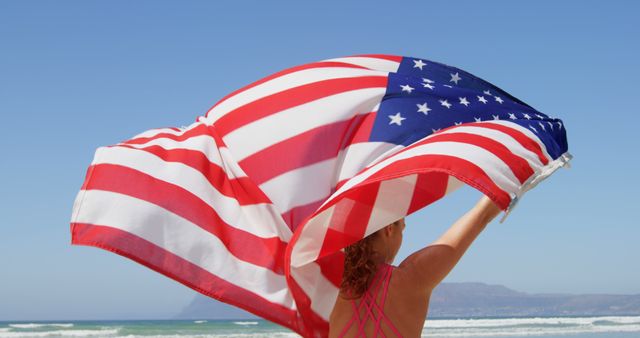 A Caucasian woman holds an American flag high on a sunny beach, with copy space. Her gesture suggests patriotism or celebration of a national holiday.