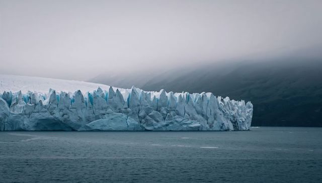 Tidewater glacier front meeting fjord under misty overcast sky, jagged seracs