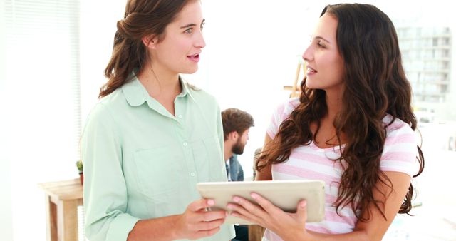 Two women engage in a work-related discussion while using a digital tablet in a modern office environment. This visual represents collaboration, teamwork, and professional communication ideal for business websites, tech blogs, and corporate training materials. The image showcases a casual and friendly office culture, useful for illustrating themes of modern workspaces and female leadership.