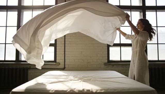 Woman throwing white sheet over bed in sunlit industrial loft with exposed brick