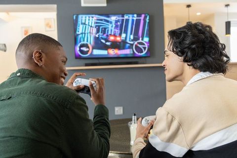 Diverse male friends playing console racing game on wall-mounted tv in open-plan lounge