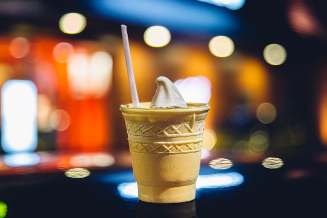 An ice cream cone-shaped cup holding soft serve ice cream with a straw, captured against colorful blurred lights in the background. This festive and playful scene can be used for food blogs, cafe marketing, dairy product advertisements, or social media posts promoting fun dessert experiences.