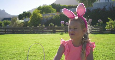Girl wearing pink bunny ears enjoying sunny backyard easter egg hunt with wicker basket