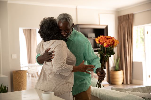 Senior African American couple embracing in their home, with the man holding a bouquet of flowers. This image can be used to depict themes of love, affection, and togetherness among elderly couples, especially during quarantine or lockdown periods. Ideal for use in articles or advertisements related to senior living, relationships, health and wellness, or pandemic-related content.