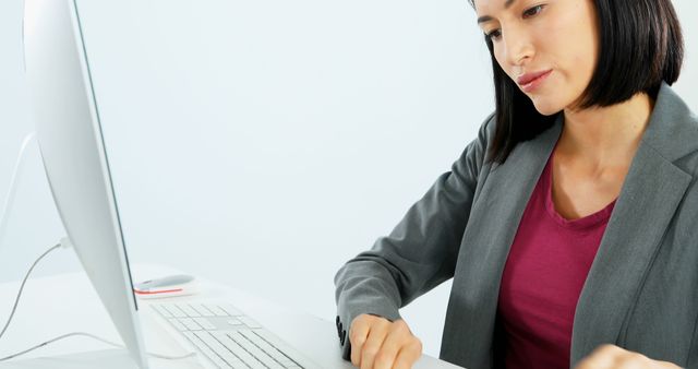 A young Asian businesswoman is focused on her work at a computer, with copy space. Her professional attire and concentration suggest a corporate or administrative environment.