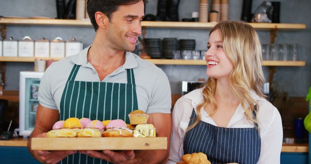 Two bakery staff members standing behind a counter, holding a wooden tray of assorted pastries and a basket of bread while smiling at each other. They both wear aprons, suggesting a professional baking environment. Ideal for content about delicious baked goods, small business teamwork and friendly customer service interactions. Suitable for promoting bakeries, cafes or baking equipment.