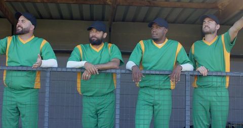 Diverse baseball teammates leaning on dugout railing watching game in green uniforms