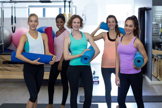 Group of diverse women holding yoga mats in a gym, smiling and ready for a workout. Ideal for promoting fitness classes, healthy lifestyle, wellness programs, and gym memberships.