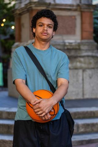 Young man standing with basketball in urban plaza wearing teal tee and crossbody bag