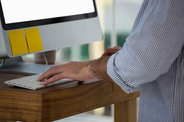 Midsection view of a designer using a computer while standing at a wooden desk in a studio. This image is ideal for illustrating modern work environments, creative professions, and ergonomic workspaces. It can be used in articles about office design, productivity, and technology in the workplace.