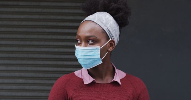 African-American woman standing outdoors wearing a protective blue face mask and headscarf in an urban setting. Ideal for content related to health, safety, pandemic measures, or public awareness about face mask usage.
