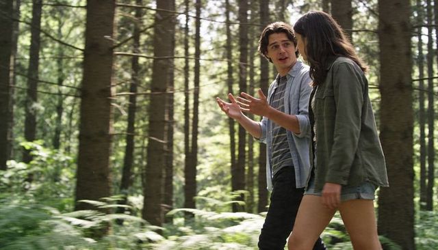 Young couple walking and chatting through sunlit conifer forest trail in casual summer outfits