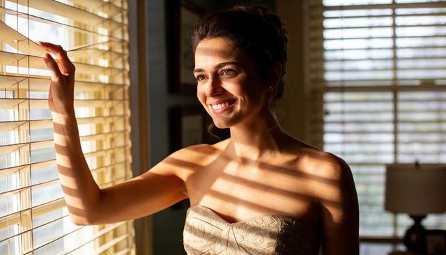 Smiling woman in strapless gown holding window blinds bathed in warm sunset stripes
