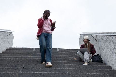 Diverse female students on campus steps using laptop and phone during coffee study break