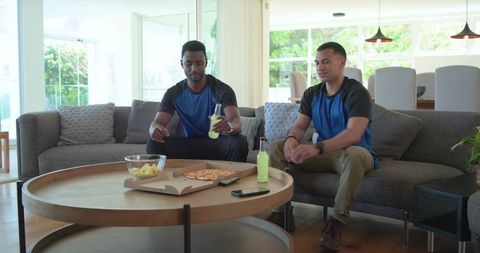 Diverse friends relaxing on sofa with pizza and drinks in bright open-plan living room