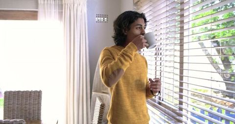Asian man standing by window drinking coffee in mustard sweater morning light interior