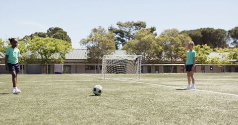 Diverse schoolgirls standing by soccer ball and goal on sunlit school sports field