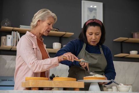 Senior women mentoring while icing layered cakes on turntable in cozy rustic kitchen