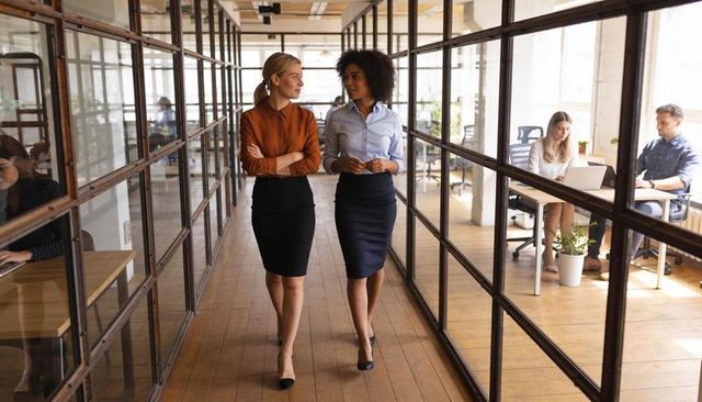 Businesswomen walking and conversing in open-plan glass office corridor for teamwork