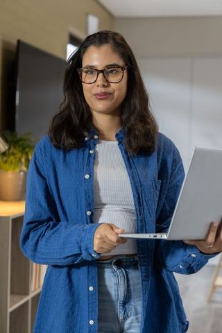 Young woman holding laptop in living room working remotely wearing denim shirt and glasses