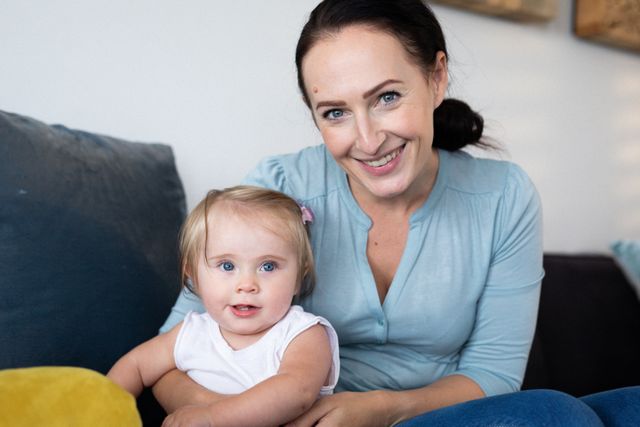 A happy mother and her baby daughter are sitting on a couch at home, smiling at the camera. This image can be used for family-oriented content, parenting blogs, advertisements for home products, or articles about quarantine and lockdown experiences.