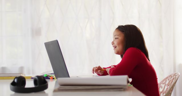 Young girl in a bright room smiling while using a laptop for online learning. Perfect for educational technology marketing, online learning platforms, homeschooling resources, and child development articles.
