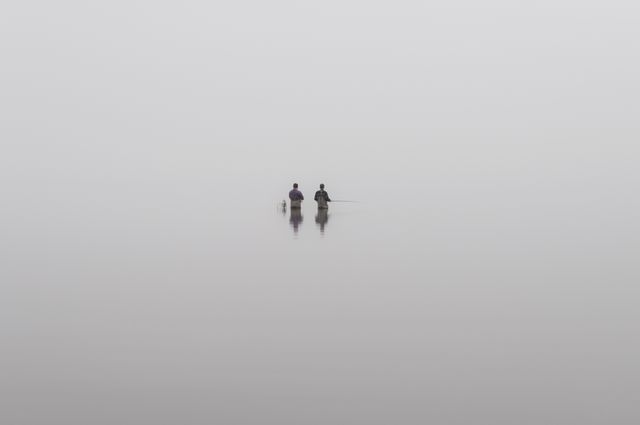 Two individuals are standing in calm, misty water, creating an atmospheric and serene scene. This minimalistic image features a tranquil reflection that can evoke feelings of peace and quiet contemplation. Ideal for concepts related to mindfulness, meditation, solitude, and peacefulness. Suitable for use in mental health awareness campaigns, nature magazines, or background themes for meditation apps.