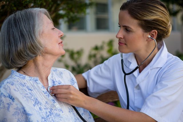 Medical professional using stethoscope to check heartbeat of senior woman in outdoor park setting. Ideal for healthcare, elderly care, medical services, and wellness promotions.