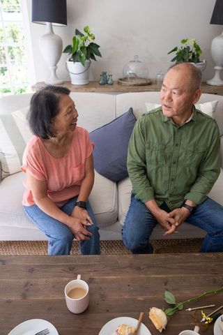 Asian senior couple sitting on sofa sharing conversation over tea in sunlit living room