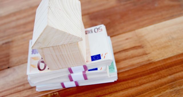 A small wooden house model sits atop a stack of Euro banknotes, symbolizing real estate investment or savings, with copy space. It represents the concept of housing market, property value, or financial planning related to home ownership.