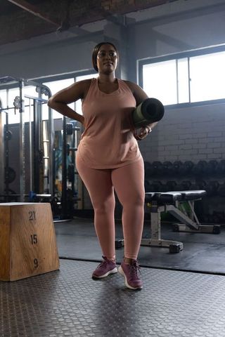 Confident african american woman standing in gym holding yoga mat wearing salmon workout