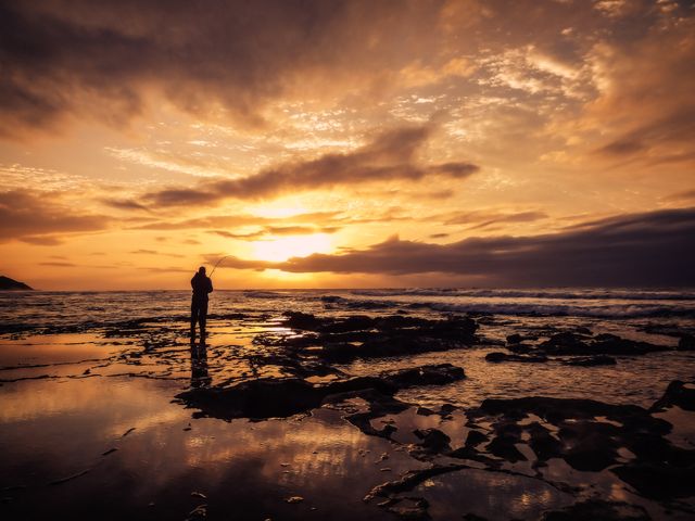 Person standing on rocky beach during sunset with dramatic clouds and sky. Reflective water on rocks and calm ocean enhance the serene and contemplative atmosphere. Ideal for use in travel brochures, inspirational content, and outdoor adventure promotions.
