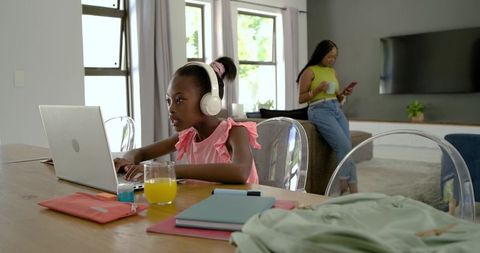 Young girl studying on laptop with headphones while parent checking phone in modern home
