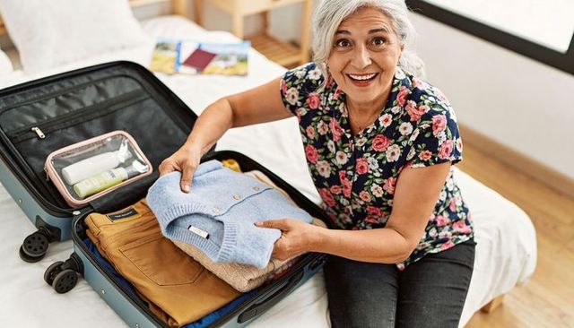 Senior woman packing suitcase at home preparing for trip with cardigan and toiletries