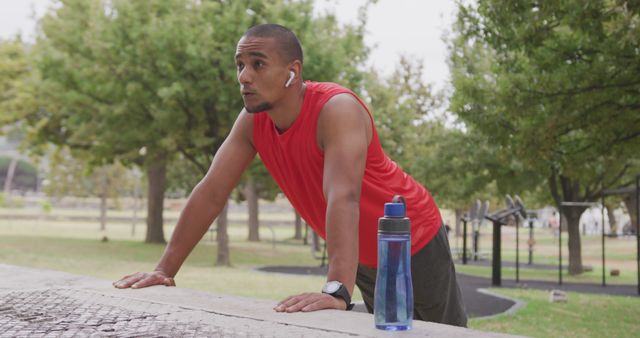 Male athlete in red sleeveless shirt resting during outdoor exercise at park. He is leaning on a structure while wearing wireless earbuds and looking focused. Blue water bottle beside him indicates importance of staying hydrated. Ideal for promoting outdoor fitness, exercise routines, and healthy living.