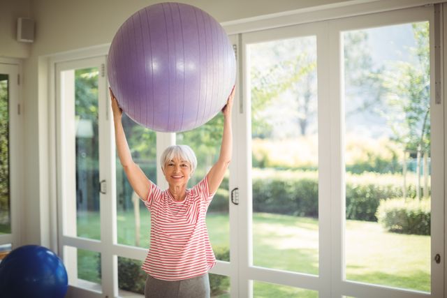 Senior woman lifting a fitness ball above her head while exercising at home. She is smiling and appears to be enjoying her workout. The room is filled with natural light from large windows, and the view outside shows a lush garden. This image can be used for promoting active lifestyles among seniors, fitness programs for the elderly, home workout routines, and healthy aging campaigns.