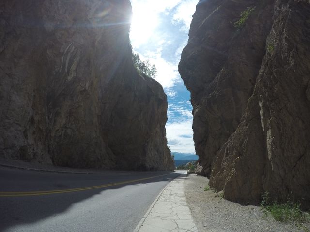 This image captures a scenic drive through a narrow mountain pass with towering rock formations on either side. The sunlight beams down, creating a striking play of light and shadow. Ideal for use in travel blogs, adventure magazines, scenic landscape collections, and promoting outdoor activities or road trips.