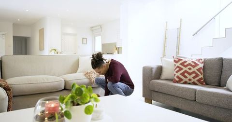 Man searching under sofa in bright modern open-plan living room with cozy decor and small plant