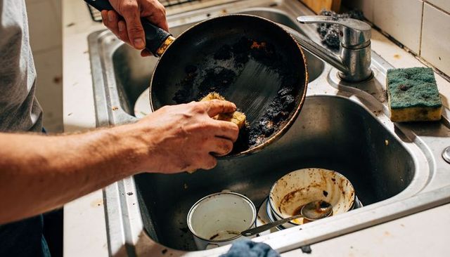 Man scrubbing burnt frying pan with sponge at messy kitchen sink, deep grease removal action