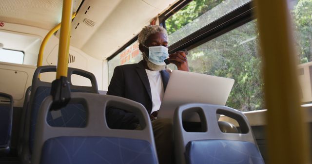 African american senior man wearing face mask talking on smartphone and using laptop while sitting in the bus. hygiene and social distancing during coronavirus covid-19 pandemic.