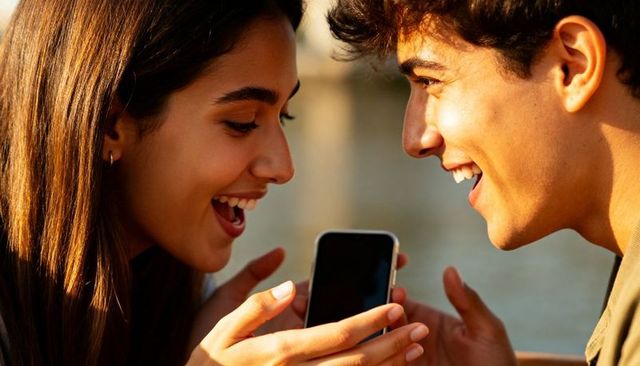 Young couple sharing smartphone, laughing during golden hour by waterfront intimate closeup