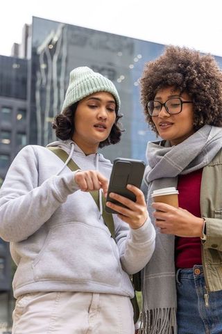 Two friends sharing smartphone and coffee on downtown street with reflective facade