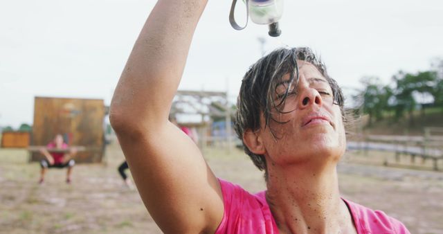 Woman pouring water on herself after a challenging mud run event. Face covered in mud, showing intense effort and determination. Great for content related to fitness, endurance, challenge, or tough outdoor activities. Perfect for articles, social media posts, and promotional materials highlighting physical resilience and perseverance.