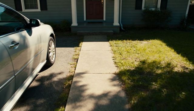 Concrete walkway leading to front porch with parked sedan and sunlit lawn