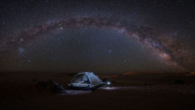 Milky way arching over desert camp with illuminated tent under starry sky