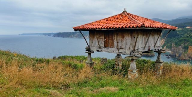 Traditional stone granary with a red tile roof stands on green grass near cliffside coastline under a cloudy sky. Historical structure shows rustic charm and antique heritage, ideal for use in travel blogs, historical documentation, cultural heritage exhibits, and coastal tourism promotions.